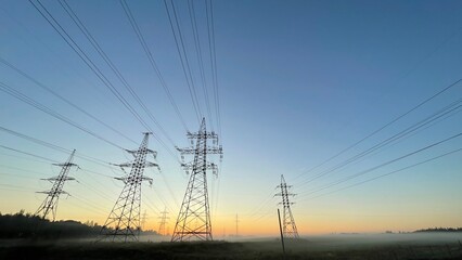 Fototapeta premium High voltage power transmission lines at sunrise with morning fog, electricity pylons across rural landscape, energy infrastructure, scenic industrial background