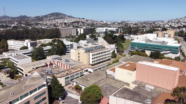 Cinematic drone shot slowly flying over the modern buildings and green spaces of the San Francisco State University campus on a bright and sunny day, with the city hills in the background