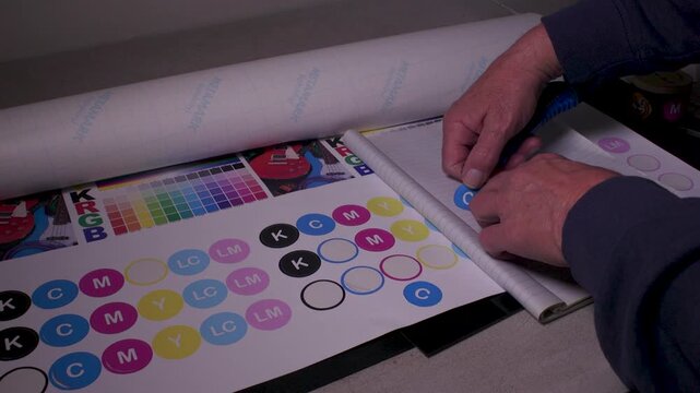 Hands use a utility knife to cut round CMYK label stickers from a calibration sheet and place them neatly onto lined paper.