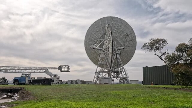 Massive ground satellite dish stands near a parked service truck on a grassy field under cloudy sky.