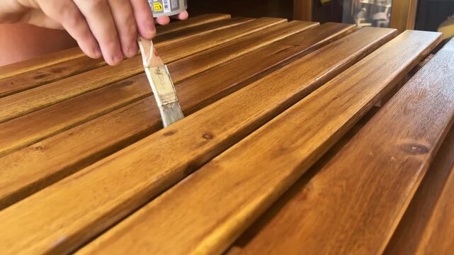 Close-up of a person carefully brushing clear varnish onto warm-toned wooden table slats in natural light.