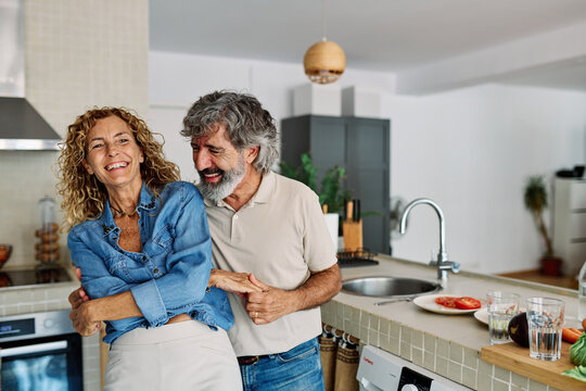 Portrait of happy senior mid aged mature couple prepering meal and having fun dancing and embracing in kitchen at hpme