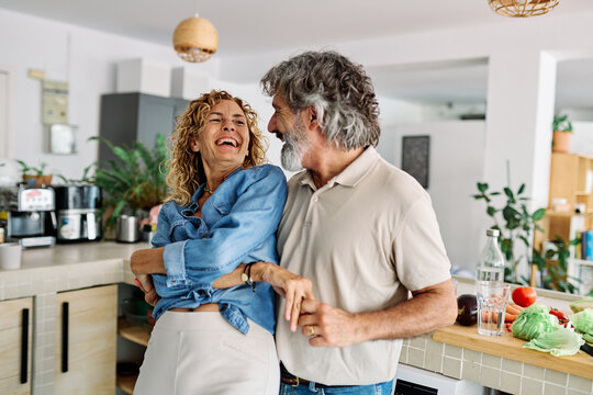 Portrait of happy senior mid aged mature couple prepering meal and having fun dancing and embracing in kitchen at hpme