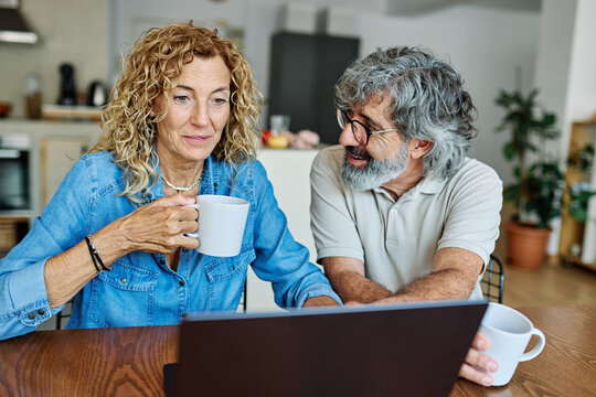 Portrait of a lovely senior mature couple using a laptop together and having fun drinking coffee or tea sitting at a table at home