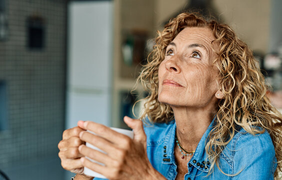 Portrait of a happy senior elderly woman holding a cup of coffee standing by window and relaxing at home