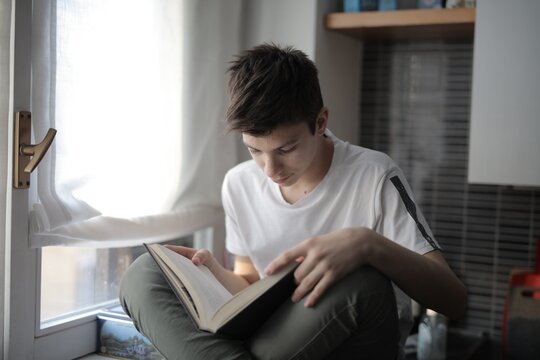 Teenager reads a book sitting on a windowsill