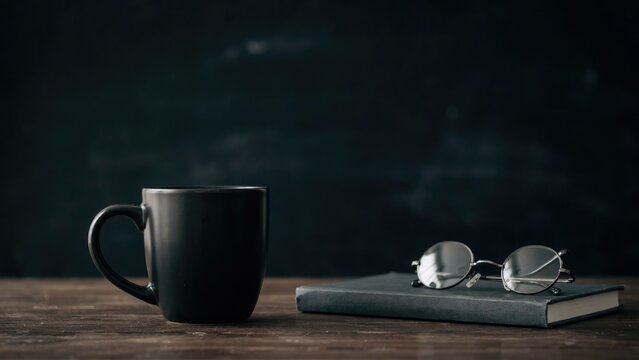 Matte black coffee mug, eyeglasses, and closed book on a dark wooden table