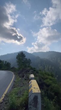 Vertical cinematic footage of a peaceful mountain road in Nathia Gali, Pakistan, surrounded by lush pine forests, rolling hills, dramatic clouds, and soft mist. 