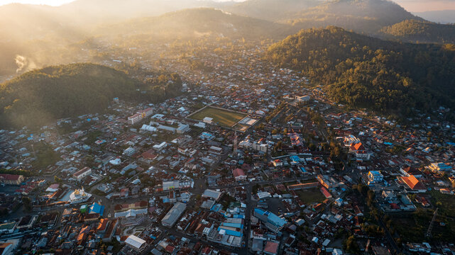 Aerial view of the town nestled among green hills at sunrise, featuring dense rooftops, a central sports field, and warm golden light in Bajawa, Nusa Tenggara Timur, Indonesia.
