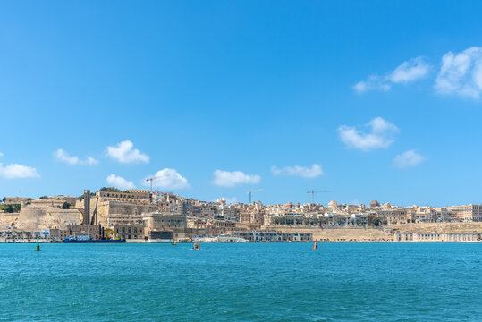 Panoramic view of Valletta, Malta, featuring historic limestone fortifications, the Grand Harbour waterfront, turquoise Mediterranean Sea, and a clear blue sky with scattered clouds