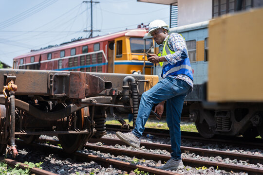 Engineer man inspecting train cars on tracks, railway engineer inspecting train carriage at logistics terminal, Railway worker using a walkie talkie to communicate during train maintenance operation