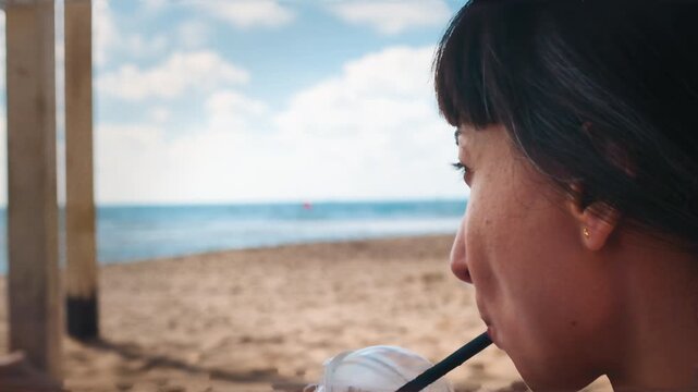 Right profile close-up of young woman sipping cold drink from clear cup with dark straw while seated on sunny sandy beach, calm coastal backdrop with blue horizon and soft light. Summer mood.
