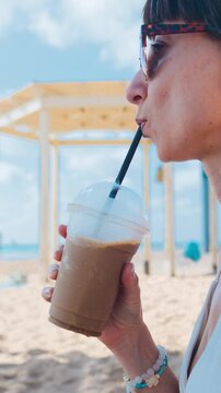 vertical video. Right profile close-up of young woman sipping cold drink from clear cup with dark straw while seated on sunny sandy beach, calm coastal backdrop with blue horizon. Summer mood.
