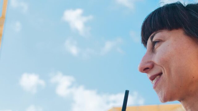 Right profile close-up of young woman sipping cold drink from clear cup with dark straw while seated on sunny sandy beach, calm coastal backdrop with blue horizon and soft light. Summer mood.