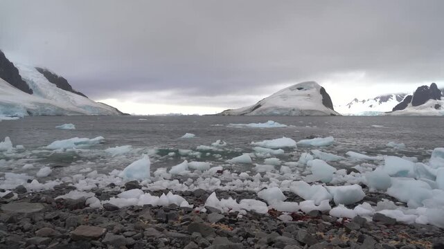 Steady wide-angle shot of blue ice chunks and brash ice washed up on the rocky shoreline of Georges Point, Rong&eacute; Island, with distant glaciers and mountains under an overcast sky
