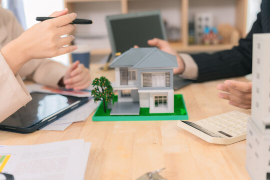 A real estate agent or architect is discussing a housing project with a client, displaying a scale model of the house and buildings alongside financial charts on the table.