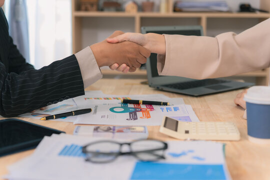 Successful business deal. Close up of business people shaking hands over desk with financial charts and laptop in office. Partnership, agreement, and real estate investment greeting concept.
