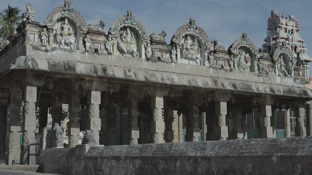 Wide shot of ancient stone mandapam with ornate Hindu deity sculptures along parapet beside temple tank. Shimmering sacred water, carved pillars, Dravidian art, Tamil Nadu, South India.