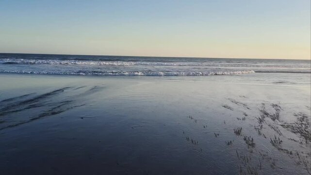 Calm ocean waves rolling onto a wet sandy beach under a clear sunset sky.
