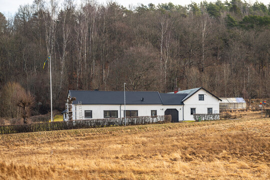 White rural farmhouse with Swedish flag beside open field and forest in quiet countryside setting.