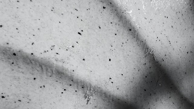 Water flows down a vertical embossed stone wall in close-up. Tiny bubbles and droplets create a moving abstract grey pattern. The minimalist relief water feature serves as modern interior decor.