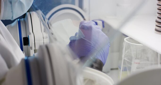 Scientist in protective gear working with equipment through glass barrier window in laboratory