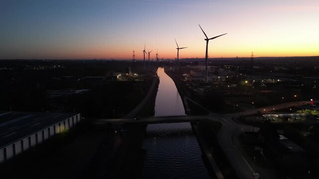 Aerial drone footage of wind turbines spinning along an industrial canal at sunset, representing renewable energy and sustainability.