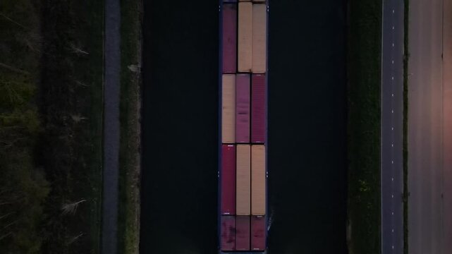 Cinematic Top-Down Aerial Tracking Shot of an Illuminated Container Ship Navigating a Canal During Twilight