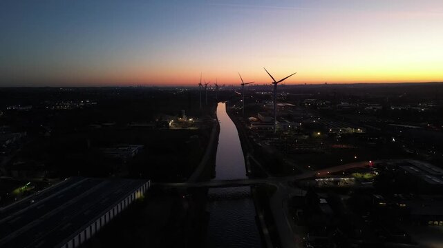 Cinematic aerial flight over an industrial canal with spinning wind turbines against a beautiful sunset sky. Sustainable energy concept.