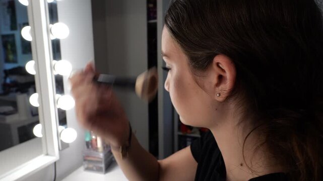 50p side view shot of a teenage girl applying makeup with a cosmetic brush in front of an illuminated vanity mirror during her beauty routine at home