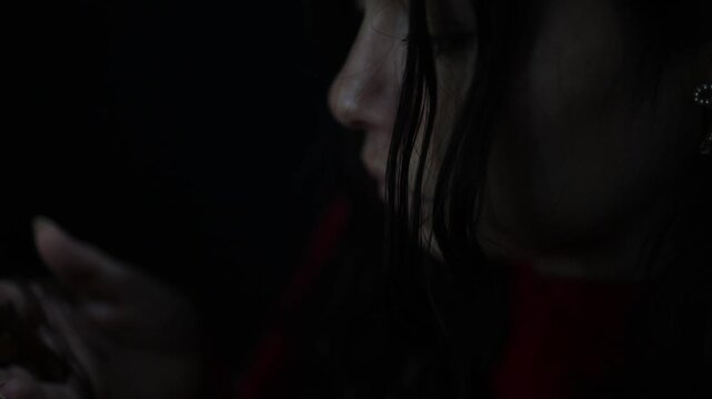 A woman in a red sweater gazes thoughtfully at cross in her hand in intimate close-up. Dramatic low key lighting creates a moody, introspective vibe. Artistic composition and shallow depth of field.
