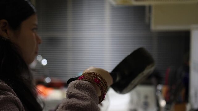 A woman in a plush sweater and bangles prepares food in a softly lit kitchen at night, using a black pan. Warm indoor lighting and shallow focus create a calm, intimate scene.