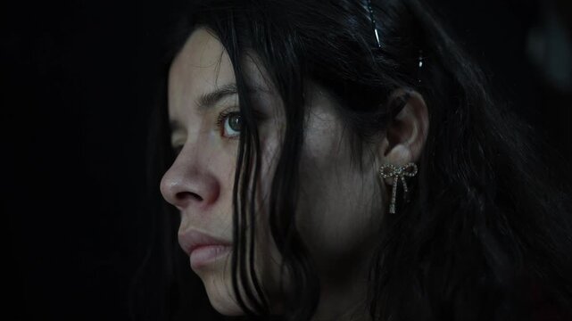 Intimate close-up shot of a Latina woman with long dark hair, silver bow earring, and contemplative expression, set against a dark background with moody soft lighting for cinematic effect.