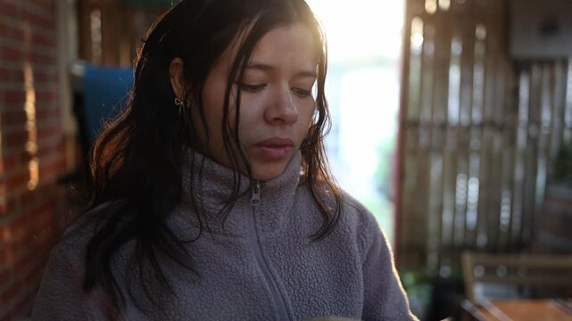 A young Latina woman in a lavender fleece jacket stands in warm golden hour sunlight, outdoors by brick and wood fencing, expressing subtle emotion. Soft natural lighting, cinematic close-up shot.