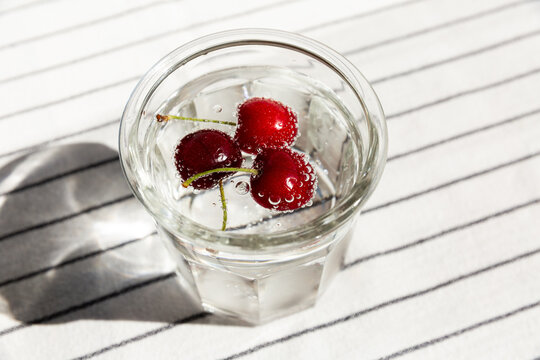 Grand verre transparent avec de l'eau p&eacute;tillante et des cerises rouges qui flottent &agrave; la surface entour&eacute;es de bulles, nappe blanche ray&eacute;e, vue du dessus, rafra&icirc;chissant