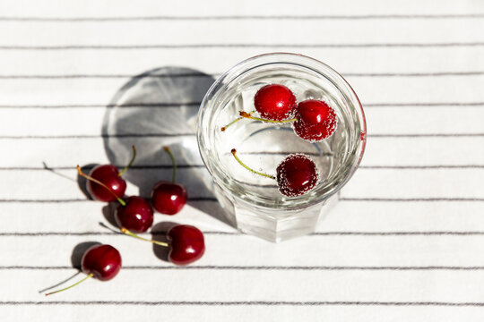 Cerises rouges qui flottent dans de l'eau p&eacute;tillante dans un grand verre transparent avec d'autres cerises pos&eacute;es sur une nappe blanche ray&eacute;e, vue du dessus, boisson d&eacute;salt&eacute;rante, bulles, &eacute;t&eacute;