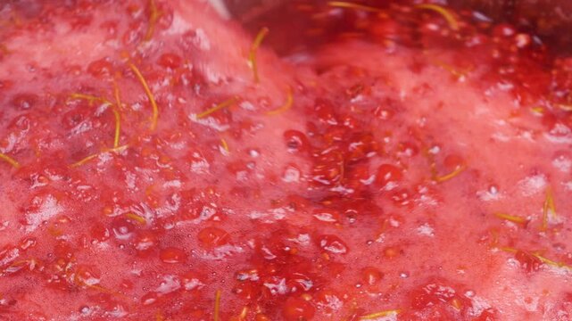 Boiling redcurrant berries in a pot to extract fresh juice for homemade jam. A chef gently stirs the hot berry mixture with a silicone spatula. The bright red mass bubbles during cooking process.