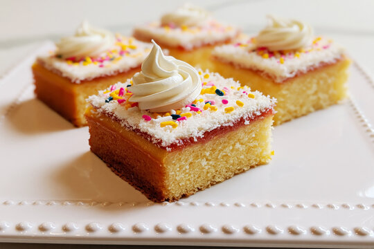 Four Petit Fours on White Plate: Four square-shaped petit fours adorned with white frosting, sprinkles, and shredded coconut sit elegantly on a white textured plate.