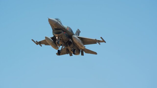 Military combat aircraft seen close from below with gear down and landing light on, fighter jet on final approach, warplane descending against blue sky to land