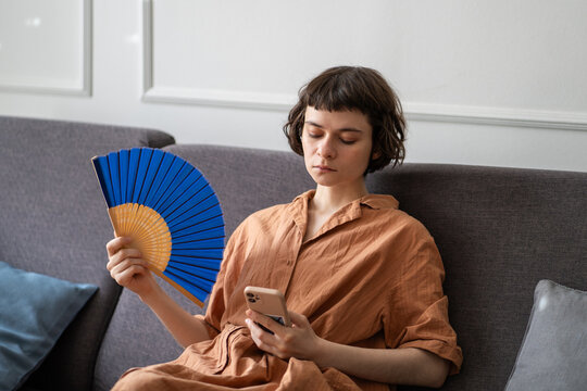 Tired woman struggling with sweltering weather using paper fan to cool down in room in summer, using mobile phone, scrolling social media. Heat exhaustion, lack of ventilation, heatwave, no ac. 