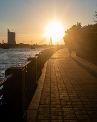 Obraz premium Golden sunset over a riverside promenade with a cable-stayed bridge, warm sun rays and a lone walking silhouette creating a peaceful urban evening atmosphere.