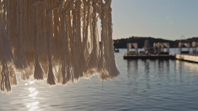 Close-up shot of white macrame fringe swaying gently with blurred luxury beach club and calm bay in golden background light, Aegean coast Turkey.