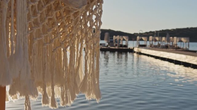 Close-up shot of white macrame fringe swaying gently with blurred luxury beach club and calm bay in golden background light, Aegean coast Turkey.