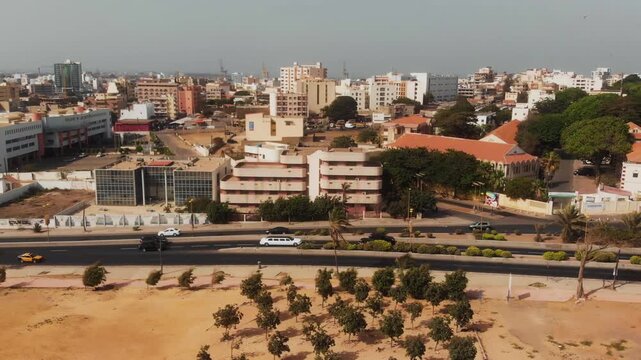Drone follows a white stretch limousine along a sandy coastal boulevard with Dakar's cityscape beyond.