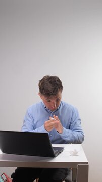 Vertical video man feeling unwell and blowing nose into a tissue while sitting at a desk with an open laptop, representing illness at work, allergies, or a common cold