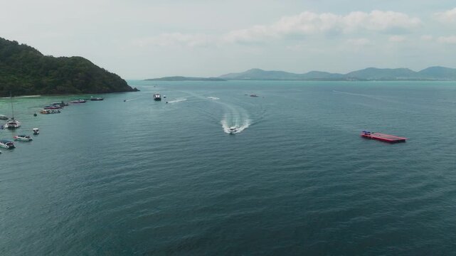 Drone pivots over a busy bay revealing boats, red platform and beach, Thailand.
