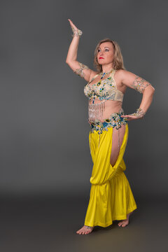 Confidence woman belly dancer with her hand raised dressing in dancing costume - bra top, yellow harem trousers and she is not wearing any footwear, performing in full length against gray background