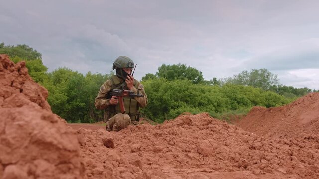 Kneeling soldier using radio and gripping rifle behind rough earthen berms in rural training field, focused on duty under cloudy overcast sky with surrounding greenery and muted natural light