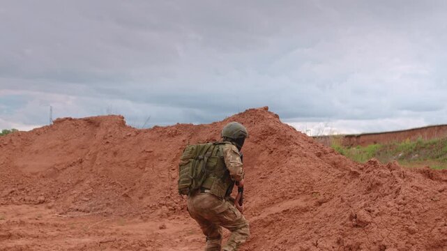 Kneeling soldier in tactical gear aims rifle across red desert ground under overcast sky, framed by earth berms, focused stance in open training terrain, with soldier, kneeling, rifle, aiming detail