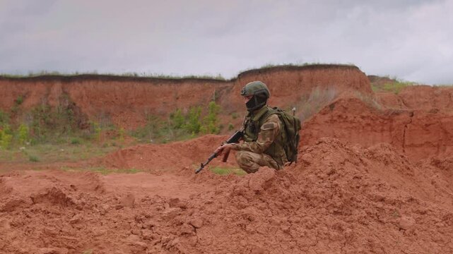 Camouflaged soldier kneeling behind red clay berm and aiming rifle while scanning open quarry landscape with helmet, face covering, backpack, radio gear, and rough terrain